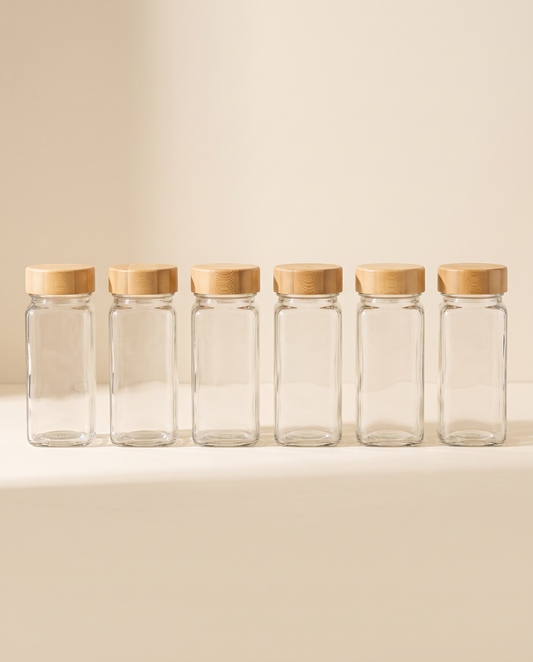 Six glass spice jars with bamboo lids on a cream background